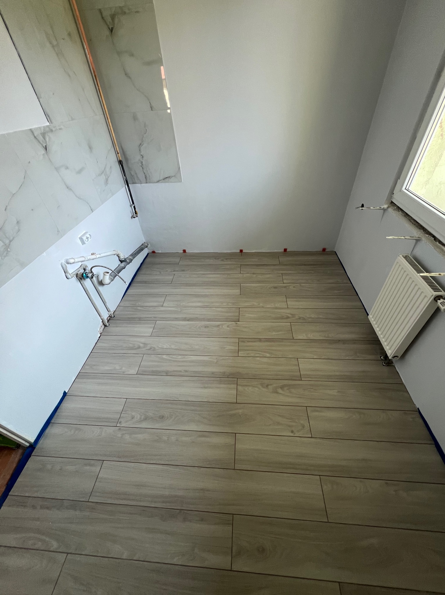 Wykończona podłoga w łazience z płytek imitujących drewno, z widocznymi rurami i grzejnikiem. Płytki na ścianie z efektem marmuru.