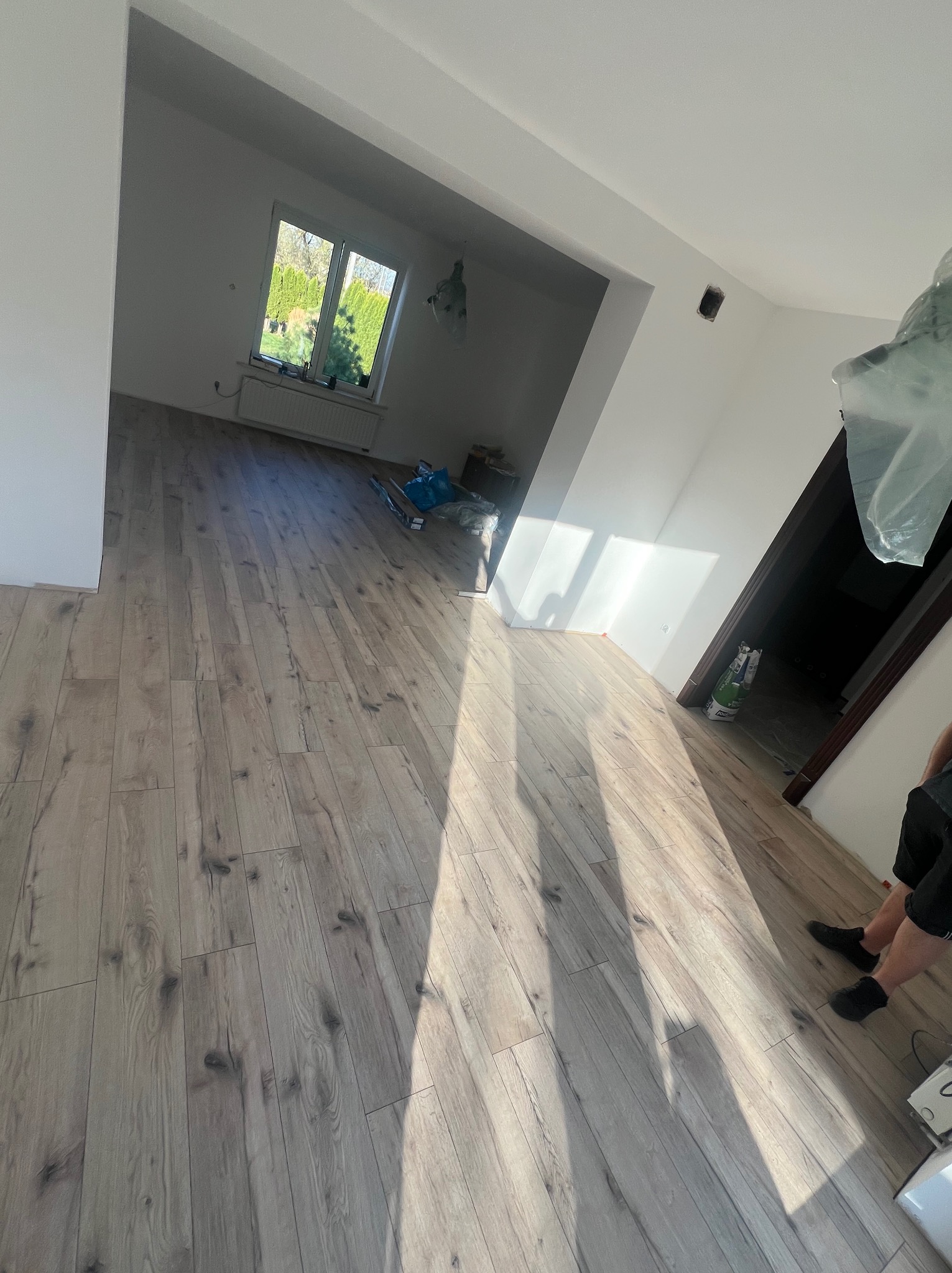 Jasne panele podłogowe ułożone w przestronnym, jasnym pokoju z oknem. Widoczne paki paneli w tle. Fragment osoby w prawym dolnym rogu kadru.