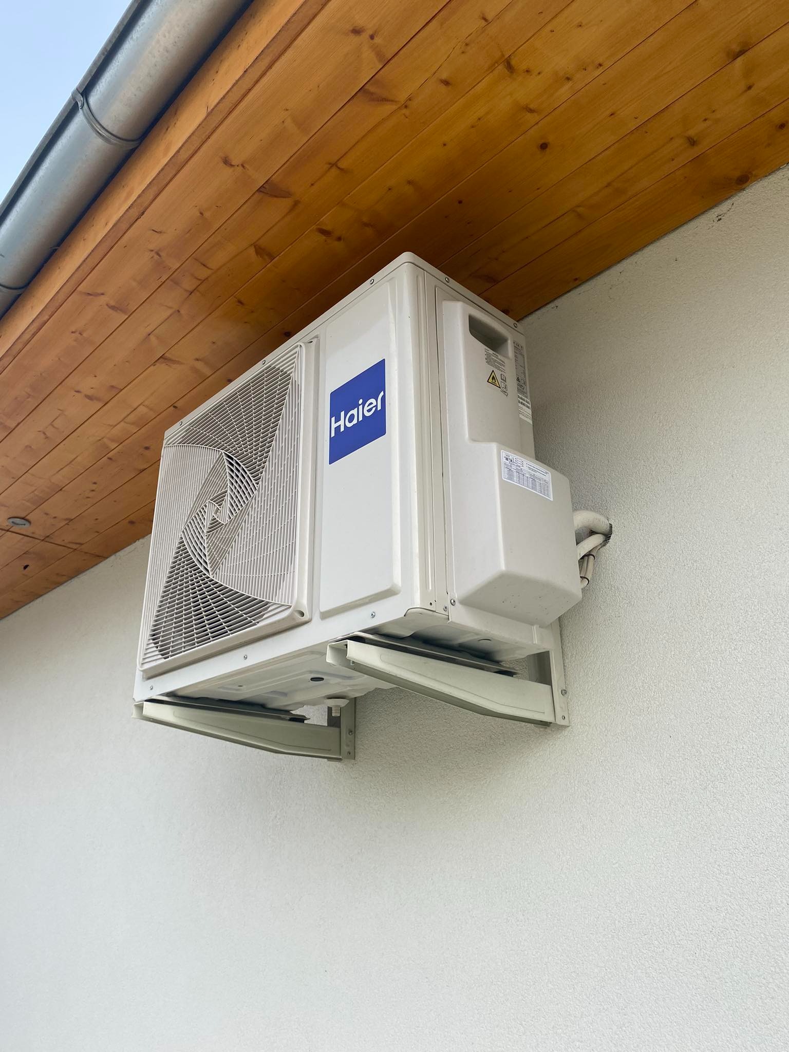Biała, zewnętrzna jednostka klimatyzacji marki Haier zamontowana na jasnej elewacji budynku z widocznym drewnianym okapem.