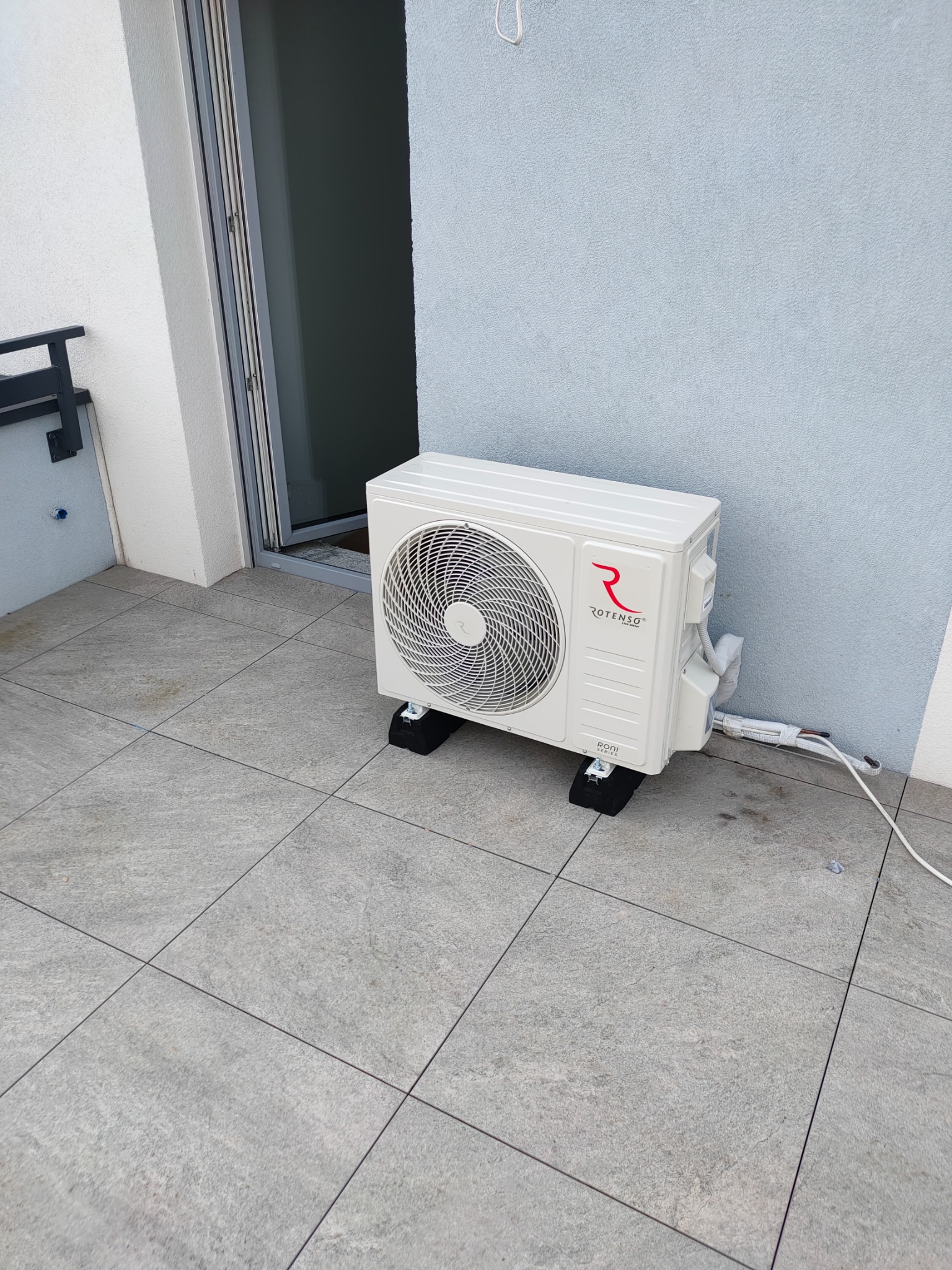 Biała, zewnętrzna jednostka klimatyzacji Rotenso umieszczona na czarnych podkładkach na szarym balkonie z płytkami, z widocznymi przewodami i fragmentem otwartych drzwi balkonowych.