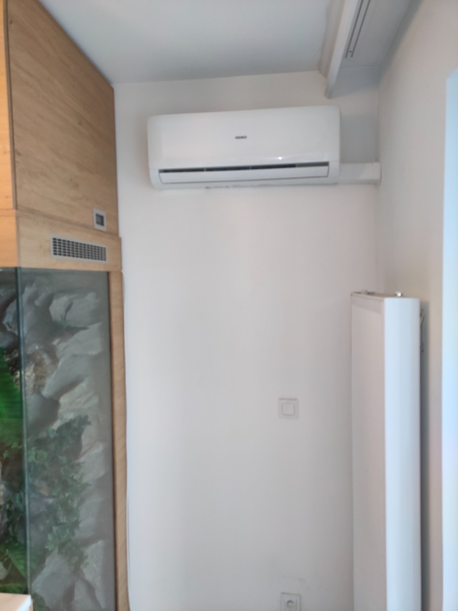 Biały klimatyzator ścienny zamontowany w pomieszczeniu z akwarium i grzejnikiem panelowym.