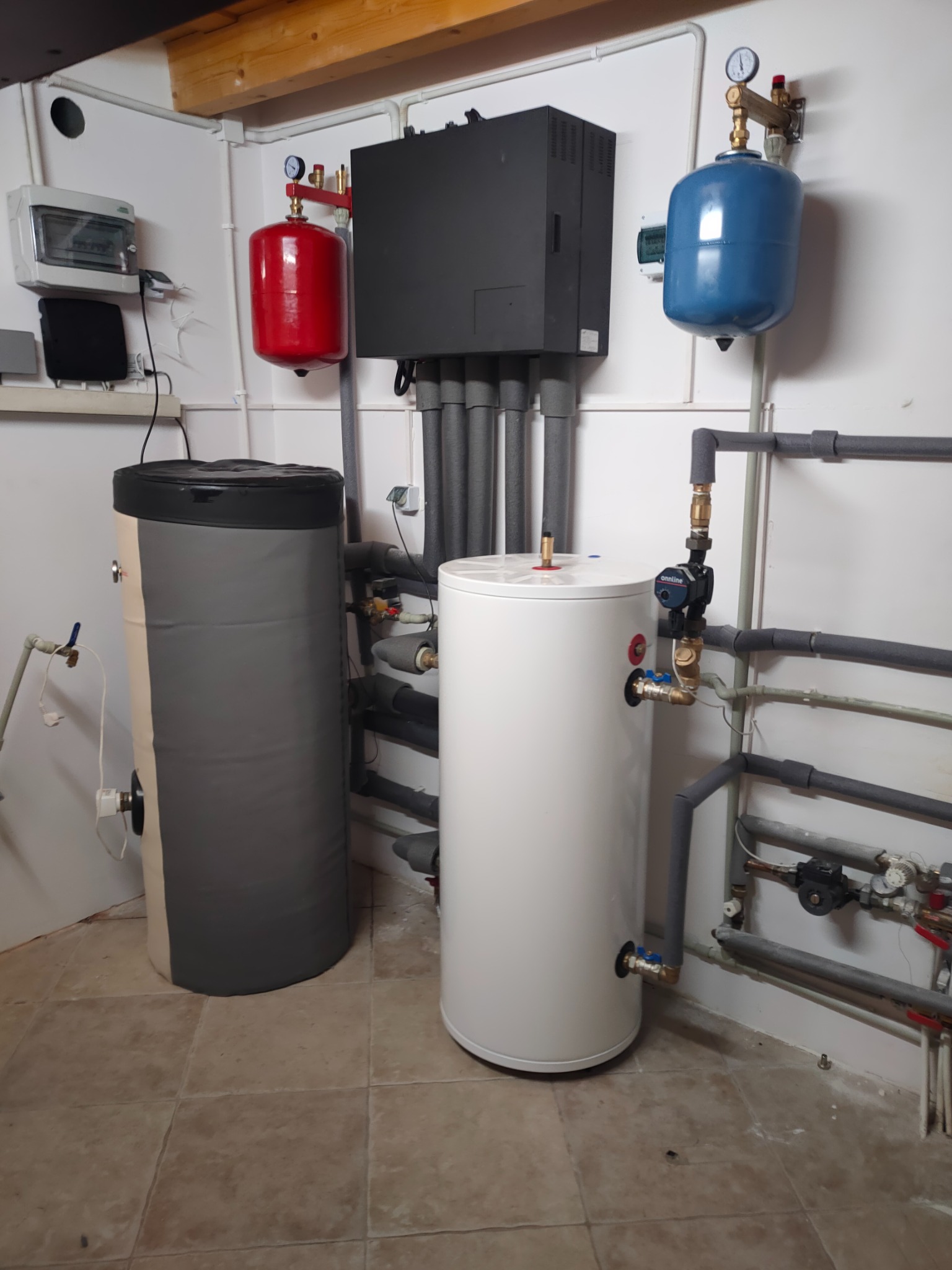 Dwa zbiorniki buforowe, szary i biały, w kotłowni z licznymi rurami i osprzętem, czerwony i niebieski zbiornik wyrównawczy ciśnienia na ścianie.