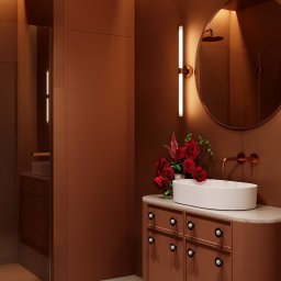 Jacek Gonera - Studio Projektowania Architektury Wnętrz - Miedziana łazienka z owalną umywalką, okrągłym lustrem i bukietem czerwonych róż. Minimalistyczny design, ciepłe oświetlenie, widoczny fragment kabiny prysznicowej.