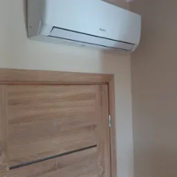 Biały klimatyzator marki Gree zamontowany nad drewnianymi drzwiami w jasnym pomieszczeniu.