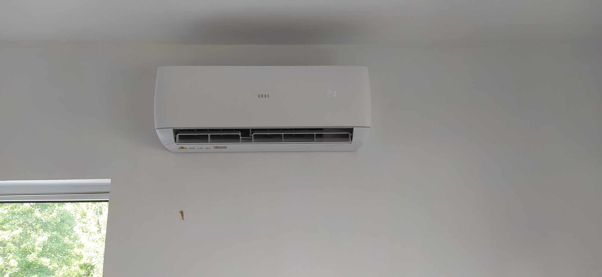 Biały klimatyzator marki ANDE zamontowany na ścianie w jasnym pomieszczeniu, widoczne okno z zielenią za szybą.