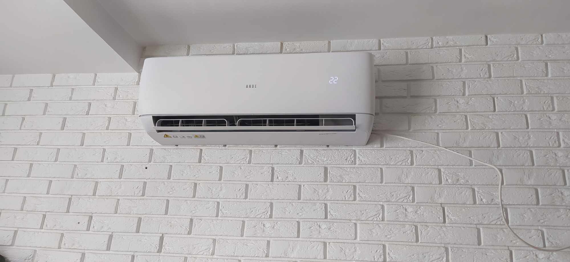 Biały klimatyzator marki ANIDE zamontowany na białej, ceglanej ścianie z wyświetlaczem wskazującym temperaturę 22 stopnie.