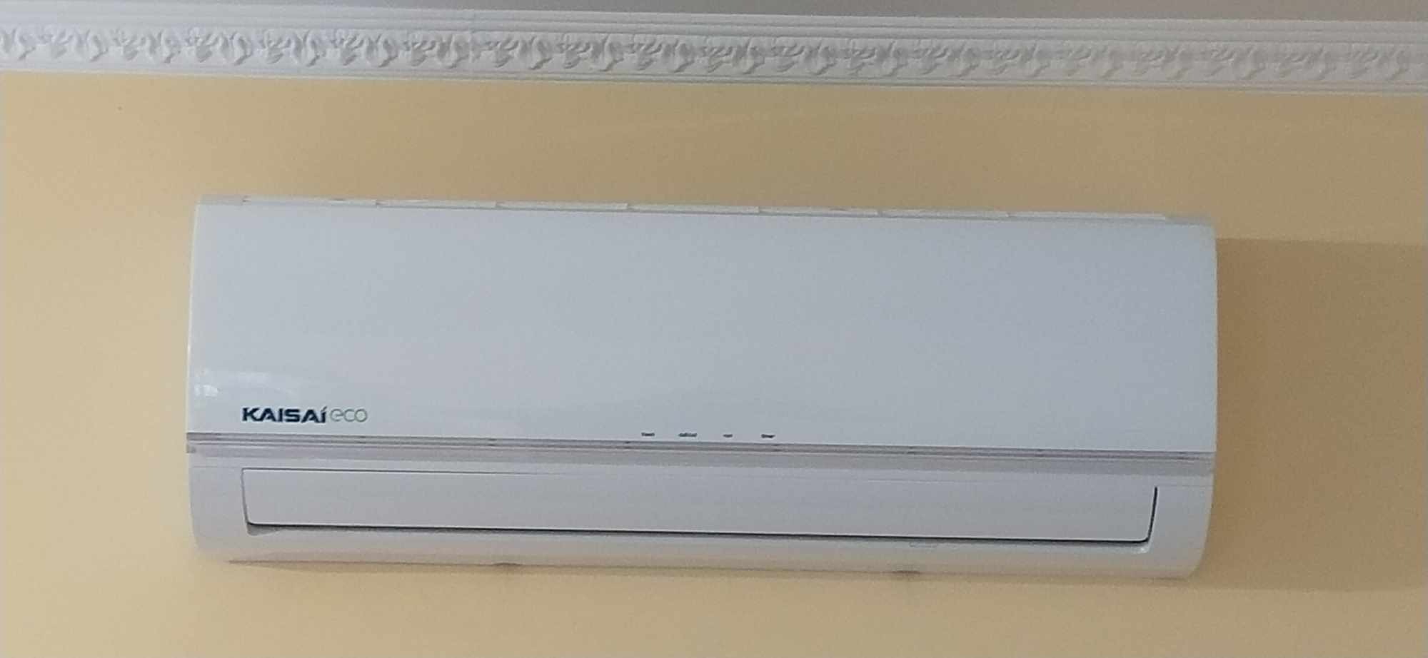 Biały klimatyzator Kaisaieco zamontowany na żółtej ścianie z dekoracyjnym gzymsem sufitowym