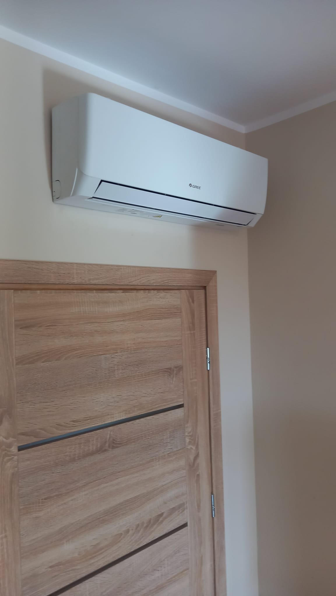Biały klimatyzator marki Gree zamontowany nad drewnianymi drzwiami w jasnym pomieszczeniu.