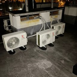 Pro Installer - Trzy klimatyzatory na dachu budynku, z widocznymi przewodami i elementami montażowymi. W tle oświetlony budynek mieszkalny nocą. Widok z góry, lekko z ukosa.