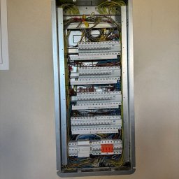 Pro Installer - Rozdzielnica elektryczna w metalowej obudowie, widoczne liczne przewody i zabezpieczenia, zamontowana na jasnej ścianie. Ujęcie z bliska, detale instalacji.
