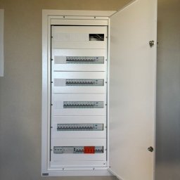 Pro Installer - Otwarta, biała skrzynka elektryczna z bezpiecznikami na ścianie o neutralnym odcieniu. Widoczne rzędy bezpieczników i okablowanie w górnej części.