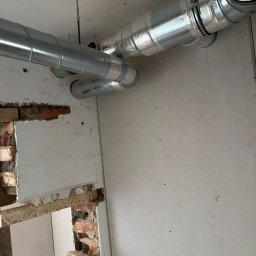 Pro Installer - Montaż srebrnych, izolowanych rur wentylacyjnych pod sufitem w surowym wnętrzu. Widoczne fragmenty cegieł i niedokończone ściany.