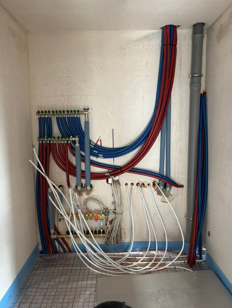 Rozdzielacz ogrzewania podłogowego z rurami PEX w kolorach czerwonym i niebieskim, ułożony na siatce grzewczej. Widoczne przewody elektryczne i rury kanalizacyjne.