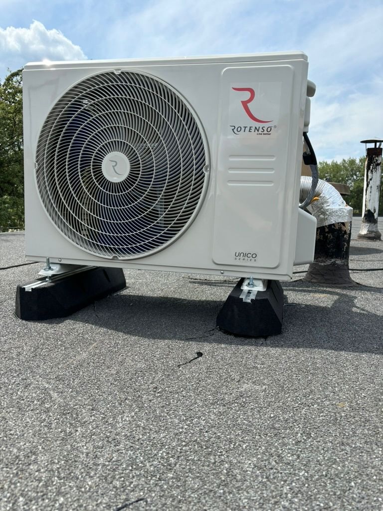 Biała jednostka zewnętrzna klimatyzacji Rotenso Unico, zamontowana na dachu z papy, z widocznym kominem w tle i błękitnym niebem.