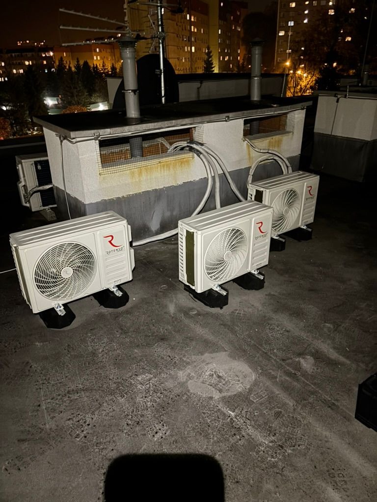 Trzy klimatyzatory na dachu budynku, z widocznymi przewodami i elementami montażowymi. W tle oświetlony budynek mieszkalny nocą. Widok z góry, lekko z ukosa.