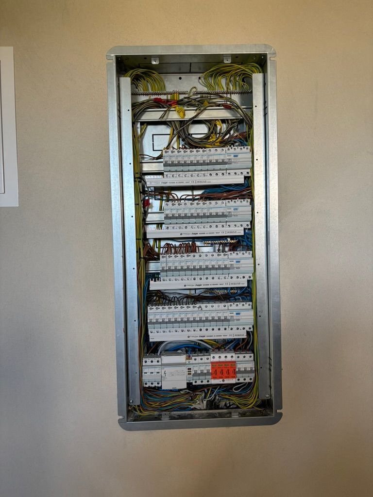 Rozdzielnica elektryczna w metalowej obudowie, widoczne liczne przewody i zabezpieczenia, zamontowana na jasnej ścianie. Ujęcie z bliska, detale instalacji.