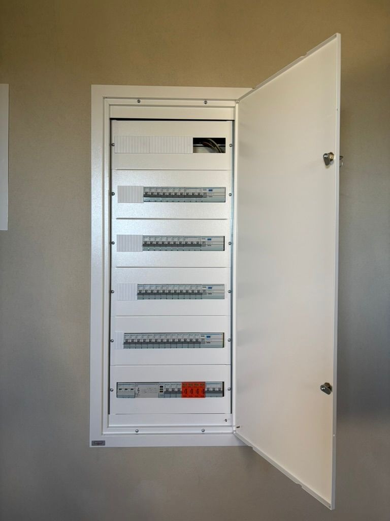 Otwarta, biała skrzynka elektryczna z bezpiecznikami na ścianie o neutralnym odcieniu. Widoczne rzędy bezpieczników i okablowanie w górnej części.