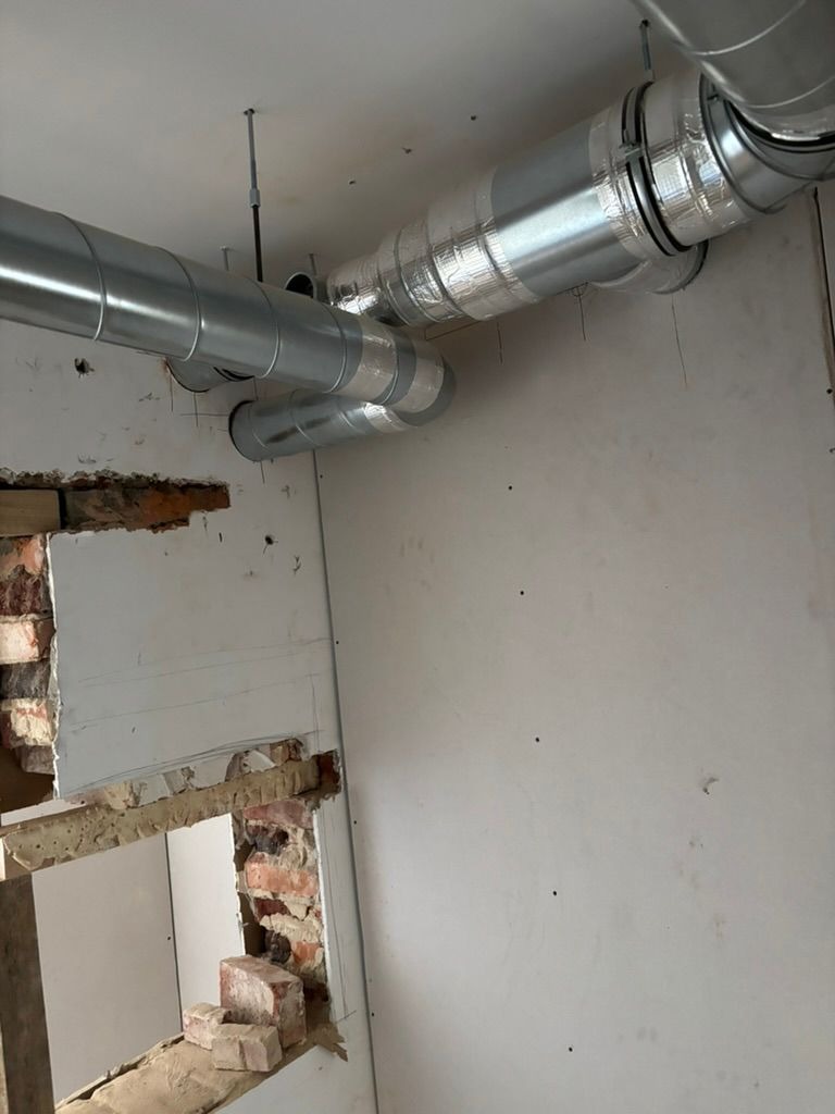 Montaż srebrnych, izolowanych rur wentylacyjnych pod sufitem w surowym wnętrzu. Widoczne fragmenty cegieł i niedokończone ściany.