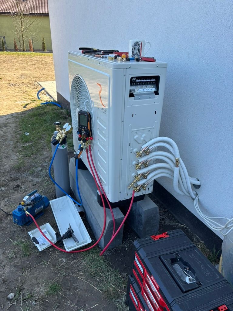 Biała pompa ciepła na betonowych blokach, z podłączonymi przewodami i narzędziami. Na górze pompy stoi kubek. Widoczne skrzynki narzędziowe i butla z gazem.