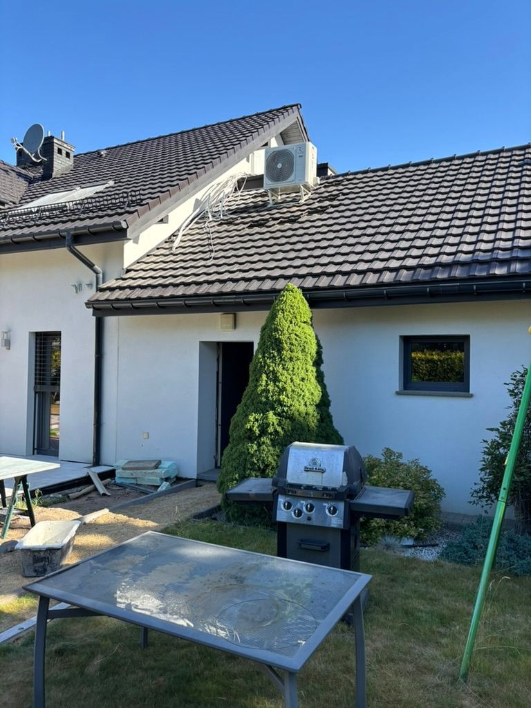 Biały klimatyzator na ciemnym dachu domu z widocznymi przewodami. Na pierwszym planie grill i stół ogrodowy, w tle zielony krzew i błękitne niebo.