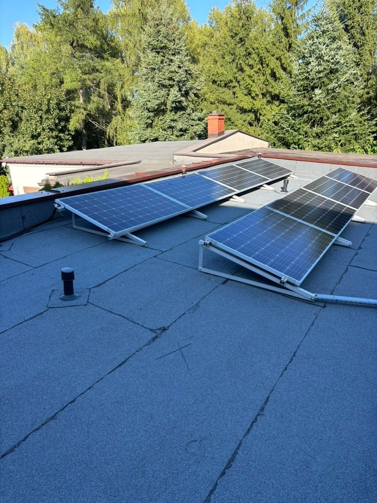 Panele fotowoltaiczne na płaskim dachu pokrytym papą, widoczne drzewa w tle. Instalacja solarna z białymi ramami, zamontowana na wspornikach. Słoneczny dzień.