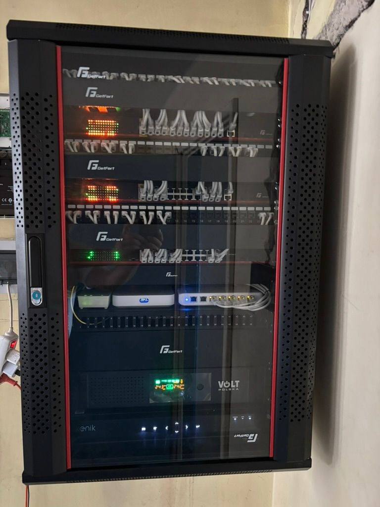 Czarna szafa rack z czerwonymi akcentami, wypełniona urządzeniami sieciowymi GetFort i Volt Polska, z widocznymi kablami i diodami LED, na tle jasnej ściany.