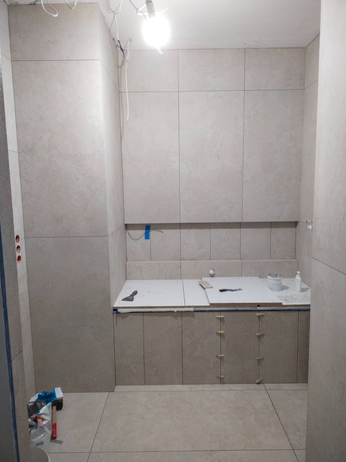 Wnętrze łazienki w trakcie wykańczania, widoczne płytki w odcieniach beżu na ścianach i podłodze, częściowo zamontowana zabudowa pod umywalkę z płytami tymczasowymi, narzędzia glazurnicze...