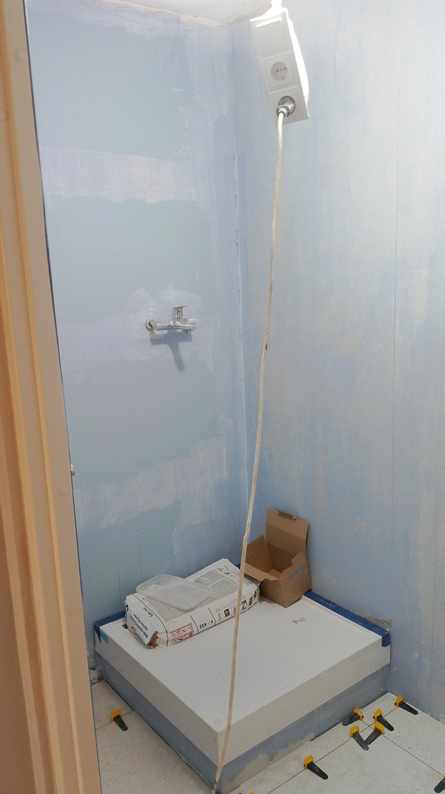 Niedokończona łazienka z przygotowanym podłożem pod prysznic, workiem zaprawy i widocznymi krzyżykami dystansowymi do płytek podłogowych; ściany w jasnym odcieniu niebieskiego, widoczne gniazdko...
