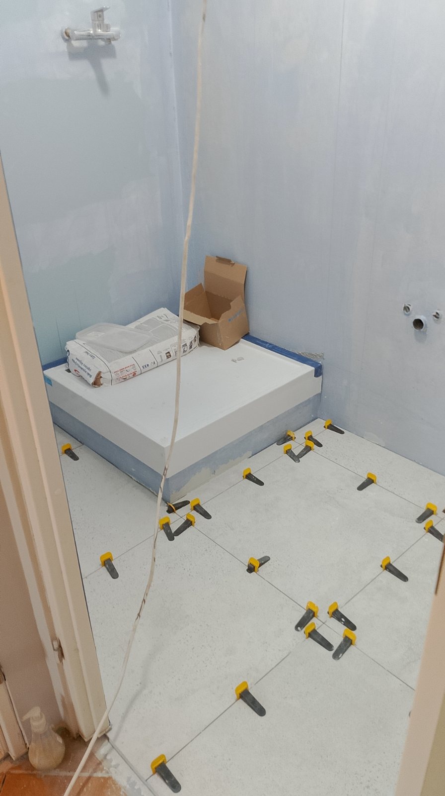 Biała posadzka w trakcie układania płytek w łazience z użyciem żółtych klinów poziomujących, widoczne worki z zaprawą i karton na podwyższeniu.