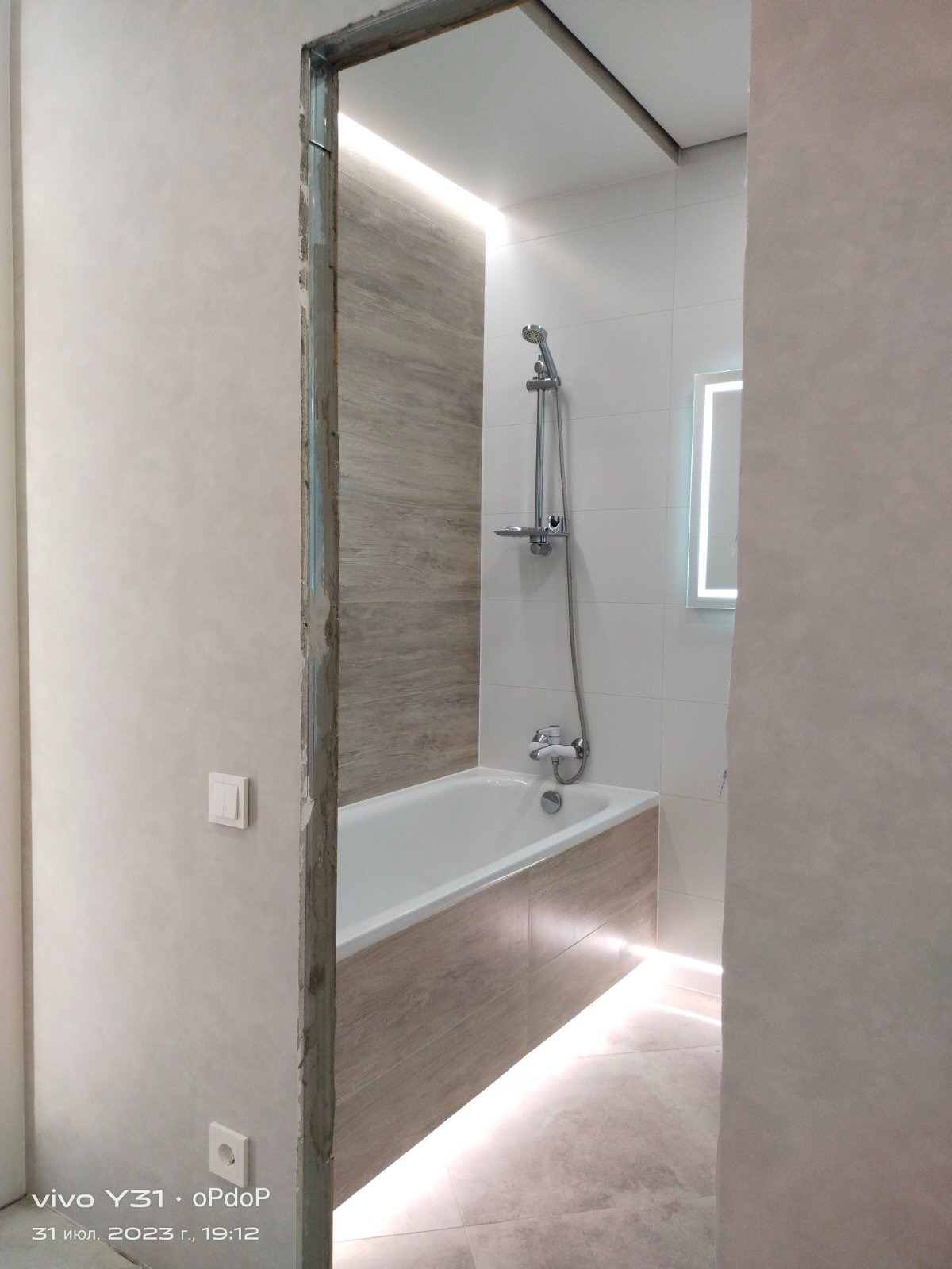 Biała wanna zabudowana panelami imitującymi drewno, chromowana bateria i prysznic, podświetlane lustro i podłoga, widok przez nieukończone obramowanie drzwi.