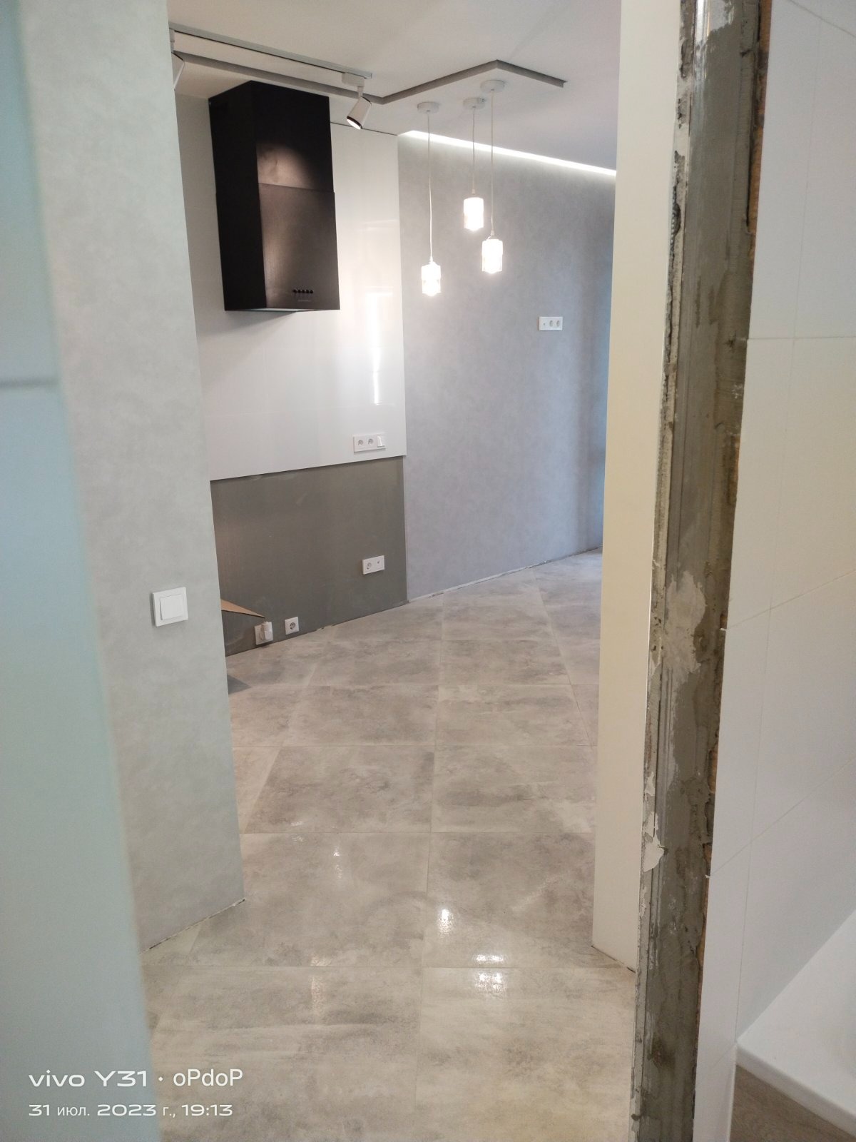 Wnętrze w trakcie wykańczania: szare płytki podłogowe, fragment ściany z widocznymi instalacjami elektrycznymi, czarny okap i wiszące lampy w tle, surowy framuga drzwi.