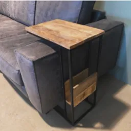 Stolik pomocniczy z drewnianym blatem i metalową ramą, umieszczony obok szarej sofy z efektem postarzenia, z półką na gazety.