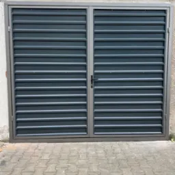 Szara, metalowa brama garażowa z poziomymi panelami, zamontowana na tle ściany z elementami starej dachówki i kostki brukowej.