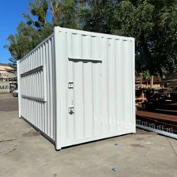 Biały, metalowy kontener biurowy z oknem i drzwiami, stojący na betonowym placu, otoczony zielenią drzew i elementami konstrukcyjnymi.