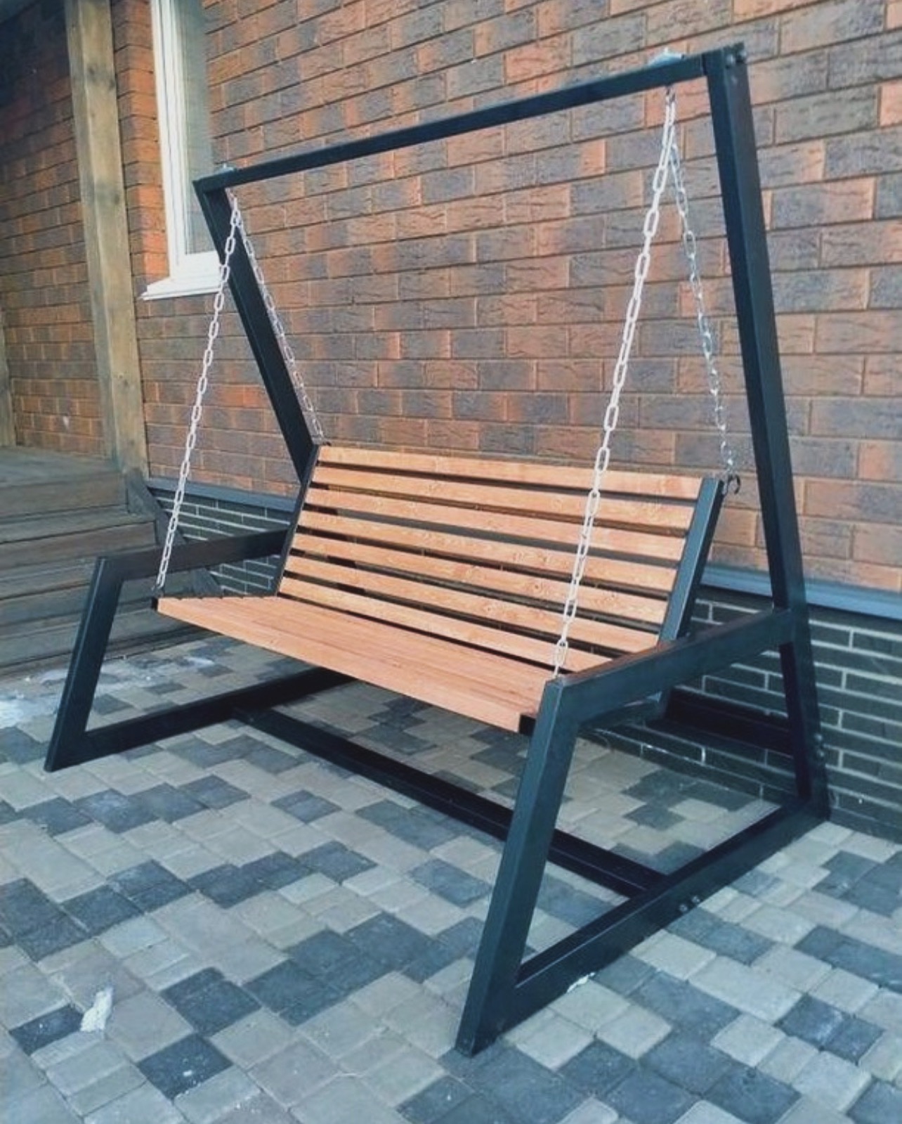 Metalowa huśtawka ogrodowa z drewnianym siedziskiem, zawieszona na łańcuchach, na tle ceglanej ściany i kostki brukowej.