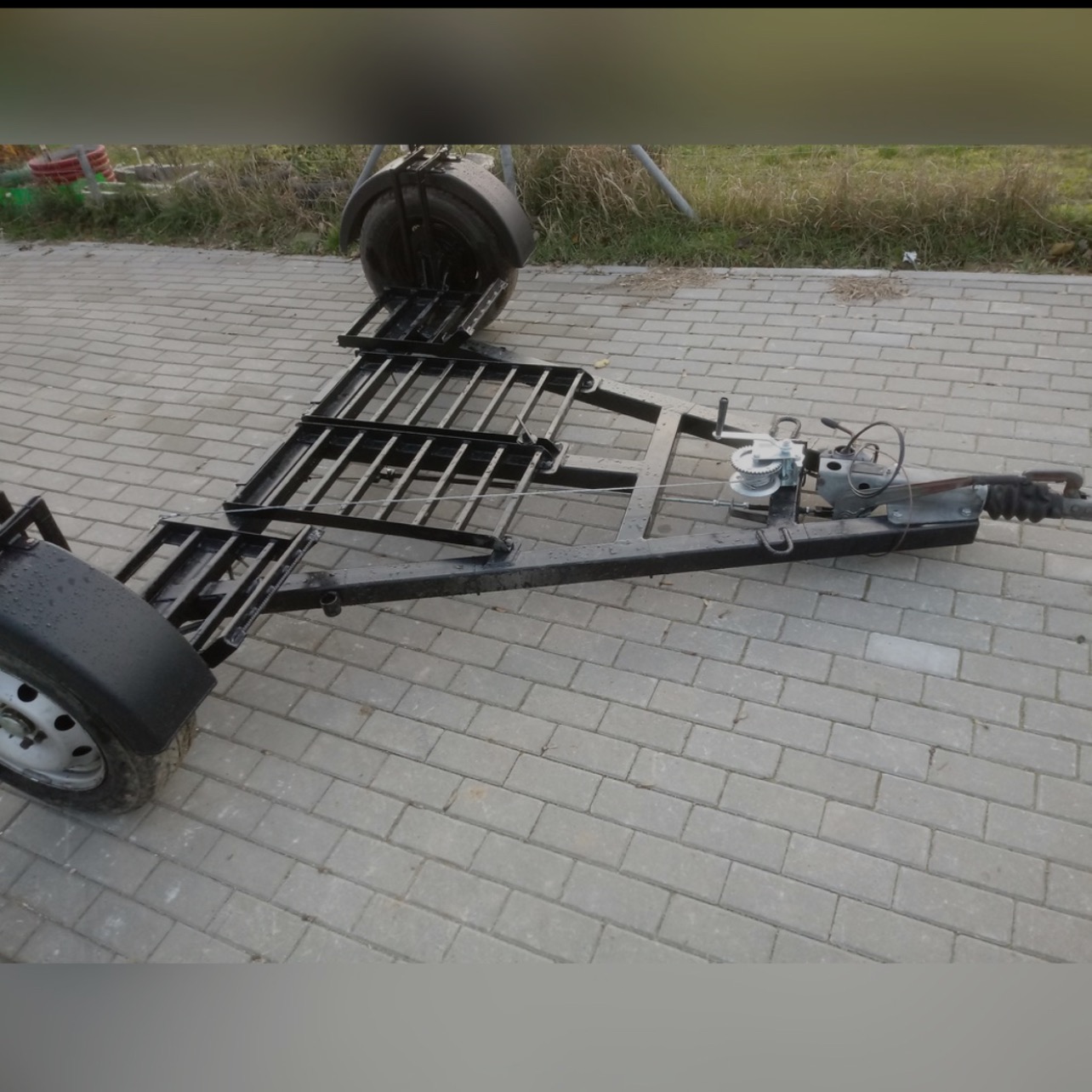 Czarna, dwuosiowa przyczepa laweta z metalową kratownicą załadunkową i ręczną wciągarką linową, stojąca na szarej kostce brukowej.