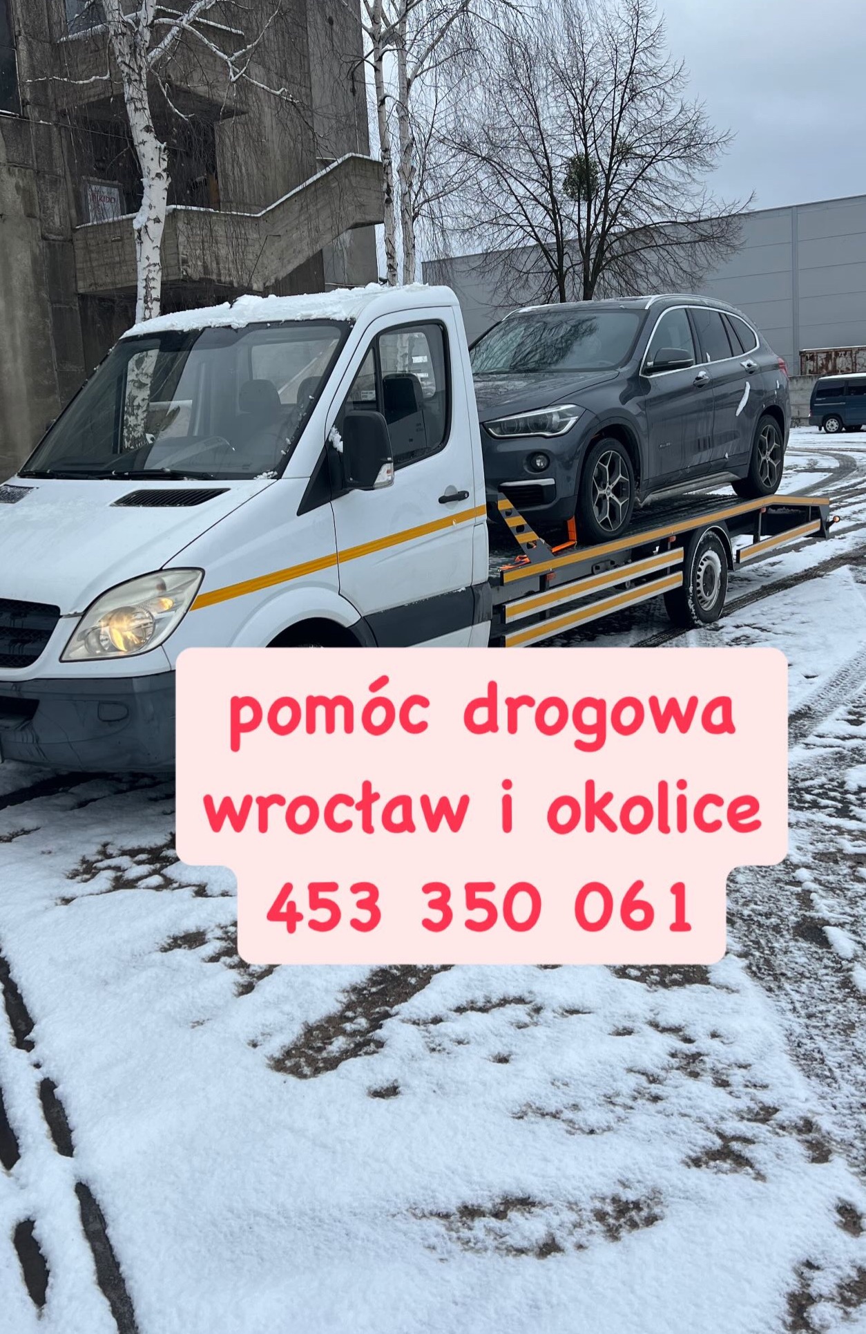 Biała autolaweta z żółtymi pasami transportuje szary samochód osobowy na tle ośnieżonego terenu i betonowego budynku.