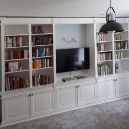 Wawrzyniak Karol - Biała zabudowa meblowa z półkami na książki, szafkami dolnymi i miejscem na telewizor, oświetlona wiszącą lampą w stylu industrialnym.