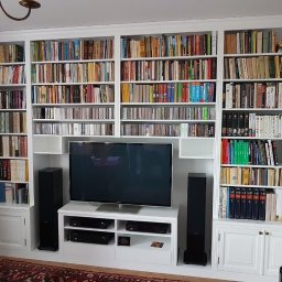 Wawrzyniak Karol - Biała zabudowa meblowa z regałami na książki i płytami CD, wkomponowana w ścianę, z szafkami dolnymi i miejscem na telewizor oraz sprzęt audio.
