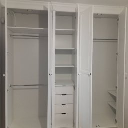 Wawrzyniak Karol - Biała, trzydrzwiowa szafa na wymiar z otwartymi drzwiami, widoczne półki, szuflady i drążki na ubrania, minimalistyczny design.