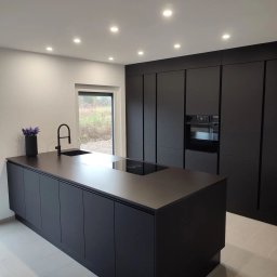 Wawrzyniak Karol - Nowoczesna kuchnia w czarnym macie z wyspą, minimalistycznym zlewem i płytą indukcyjną, oświetleniem punktowym oraz widokiem na zewnątrz przez okno.