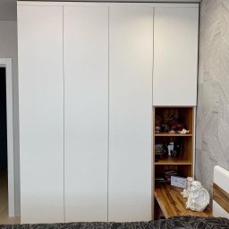 Wawrzyniak Karol - Biała szafa wnękowa z otwartą drewnianą półką, na której znajdują się dekoracyjne przedmioty i mała figurka anioła na wysuwanym blacie.