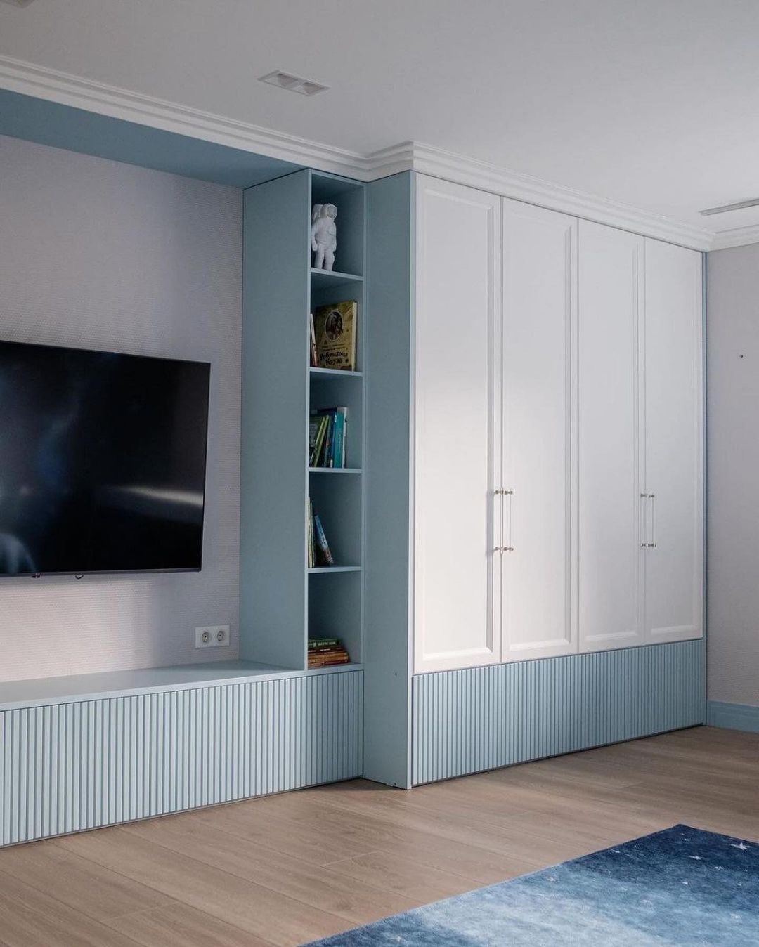 Biała szafa wnękowa z niebieskimi akcentami, połączona z szafką RTV w tym samym kolorze, z otwartymi półkami wypełnionymi książkami i figurką astronauty, na tle ściany z telewizorem.
