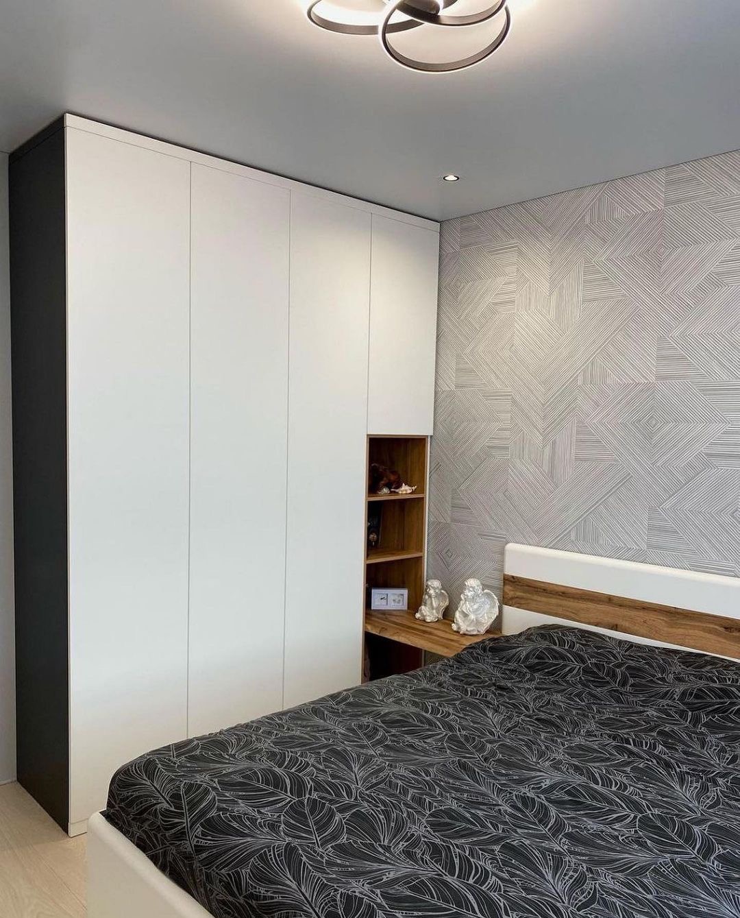 Nowoczesna sypialnia z białą szafą w zabudowie, fragmentem otwartych półek z drewnianym wykończeniem i dekoracyjną tapetą w geometryczne wzory.