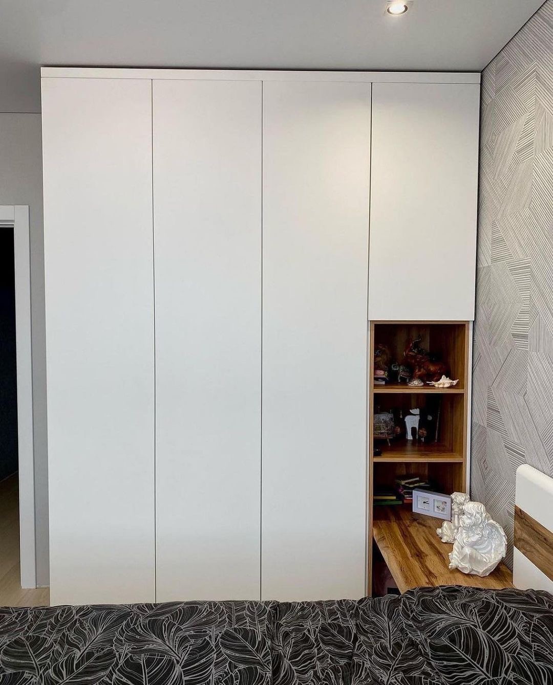 Biała szafa wnękowa z otwartą drewnianą półką, na której znajdują się dekoracyjne przedmioty i mała figurka anioła na wysuwanym blacie.