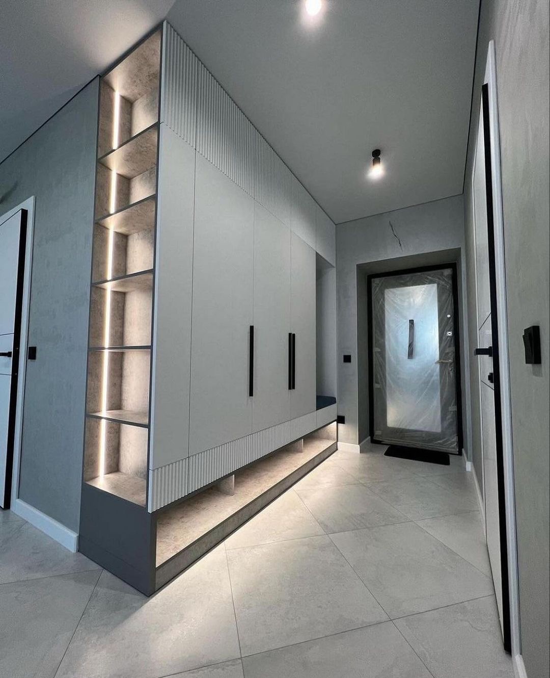 Szafa wnękowa w przedpokoju z oświetleniem LED w półkach, szare fronty i czarne uchwyty, widoczna ławka wbudowana w szafę, drzwi wejściowe zabezpieczone folią.