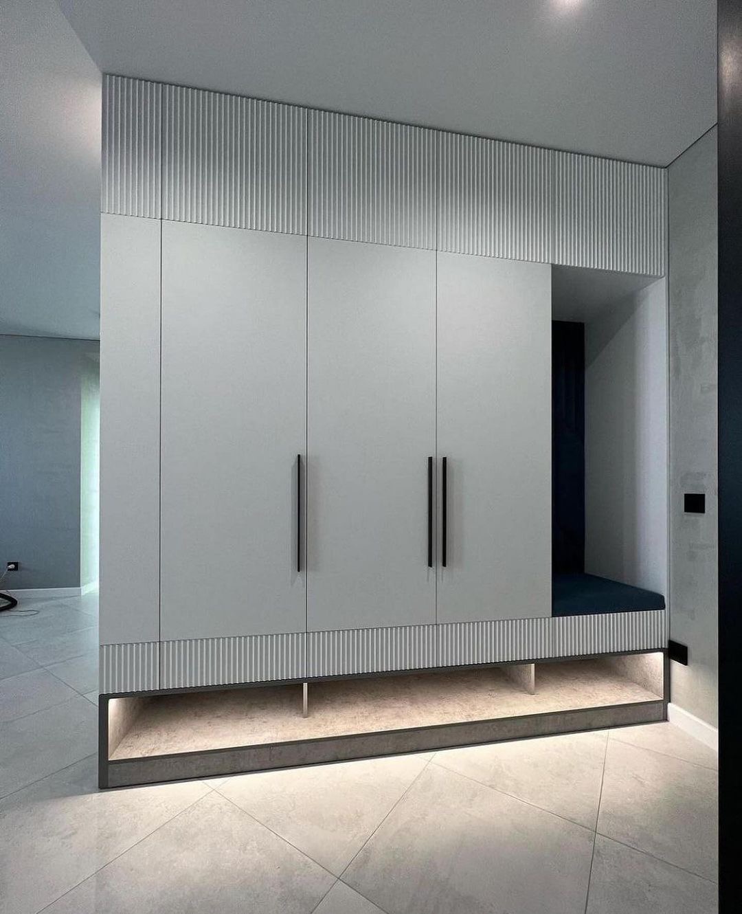 Nowoczesna zabudowa wnęki z szafą z białymi frontami i siedziskiem z granatowym obiciem, z oświetleniem pod spodem i dekoracyjnymi panelami ryflowanymi na górze i na dole.