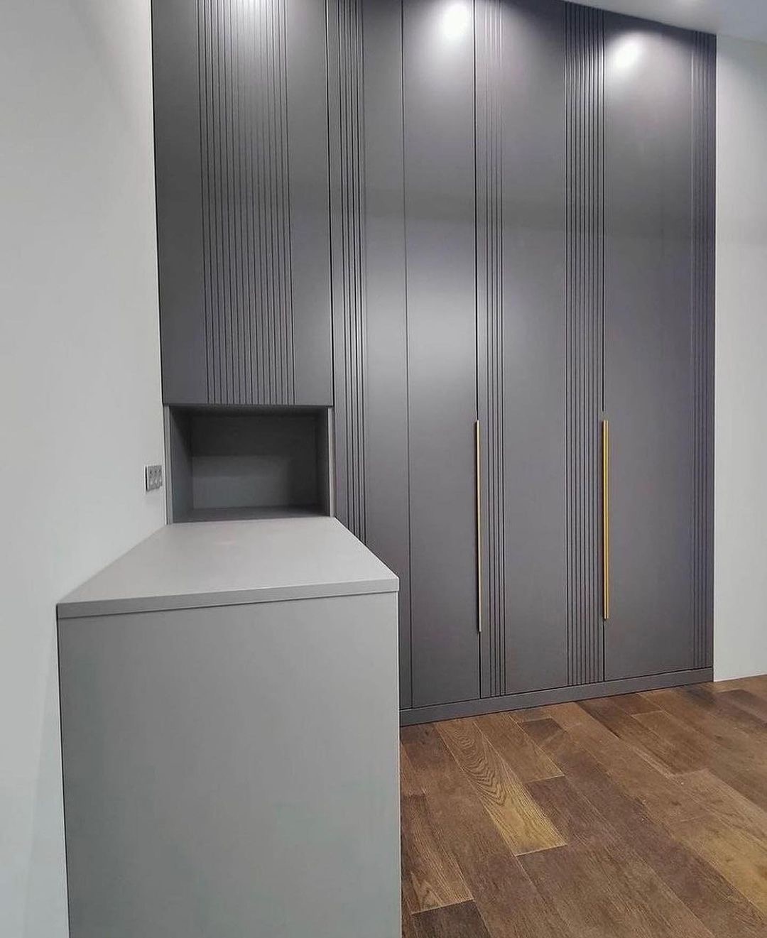 Szara zabudowa meblowa z szafami z pionowymi frezami, zintegrowana z komodą z otwartą półką, na drewnianej podłodze.