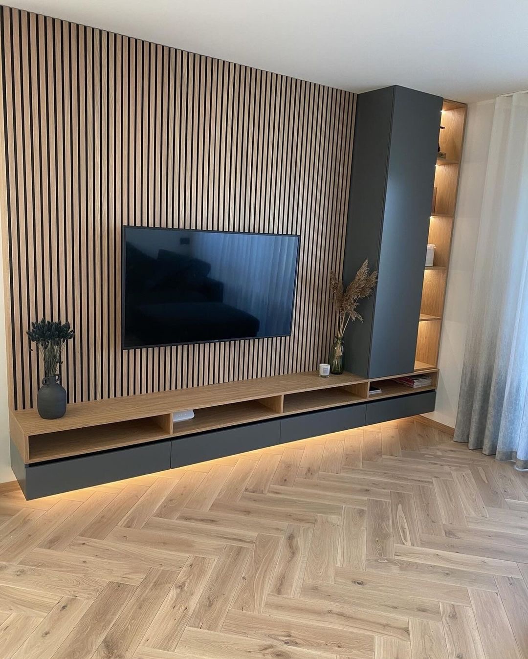 Nowoczesny salon z drewnianą ścianą z pionowych paneli, podwieszaną szafką RTV z podświetleniem LED, szarą kolumną i parkietem w jodełkę.