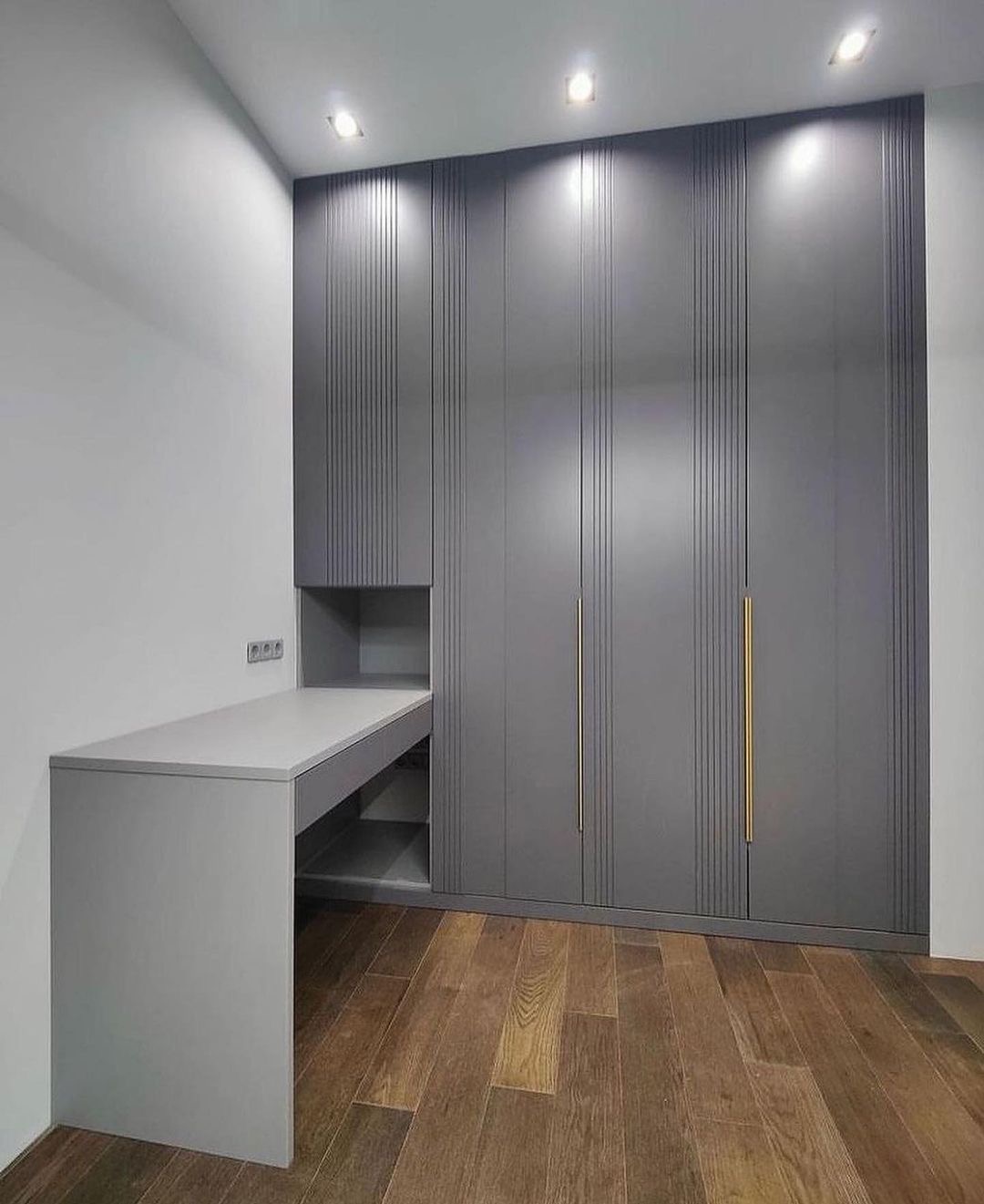 Szara szafa wnękowa z pionowymi żłobieniami i złotymi uchwytami, zintegrowana z biurkiem w kolorze, na drewnianej podłodze.